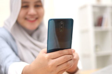 Asyalı Müslüman kadının akıllı telefon kullandığı bir fotoğraf. İnternette iletişim kurarken mutlu gülümseme hareketi, video çağrısı, seçici odaklanma 