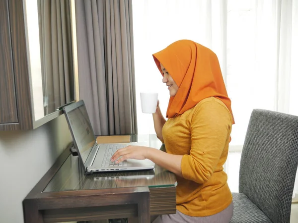 Asian muslim woman wearing hijab smiling while typing on her laptop, work from home concept. Young successful female businesswoman or entrepreneur working at home