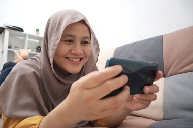 Güzel Asyalı Müslüman kadın koltukta dinlenirken, oyun oynarken ya da internetten video izlerken mutlu bir gülümseme ile gülümsüyor.