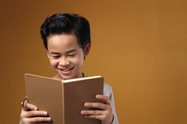 Bir kitap okurken gülümseyen Asyalı genç çocuğun portresi, sarı arka plana karşı eğitim kavramı.