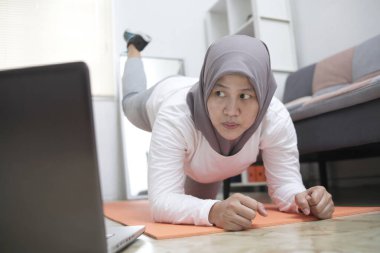 Asyalı müslüman kadın tesettür takıyor evde egzersiz yaparken dizüstü bilgisayarda video eğitimi, kapalı ev antrenmanı konsepti izliyor, yeni normal yaşam tarzında sağlıklı kalıyor.