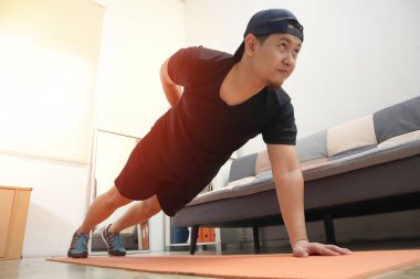 Asyalı erkek, yeni normal yaşam tarzında sağlıklı kalmak için evde egzersiz yapıyor. Ev içi antrenman konsepti, tek kolu kalas pozisyonunda.