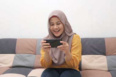 Güzel Asyalı Müslüman kadın koltukta dinlenirken, oyun oynarken ya da internetten video izlerken mutlu bir gülümseme ile gülümsüyor.