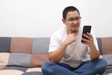 Mutlu gülümseyen Asyalı adam evdeki koltukta otururken akıllı telefon kullanıyor. Evde dinlenirken cep telefonunda mesajları okuyan bir erkek