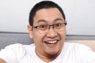 Asyalı bir adam selfie çekiyor ya da evde video görüşmesi yapıyor kameraya gülümsüyor ve el sallıyor. Mutlu neşeli ifade