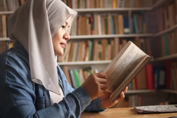 Asian muslim woman wearing hijab reading book in library, educational concept. Happy smiling expression when doing leisure activity