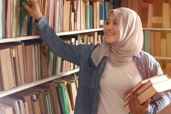 Asian muslim woman wearing hijab picking book in bookshelf, education concept, reading learning studying in library, happy smiling expression