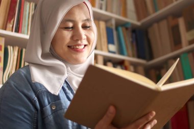 Kütüphanede tesettürlü Asyalı Müslüman kadın kitap okuyor, eğitici bir kavram. Boş zamanlarında gülen mutlu yüz ifadesi