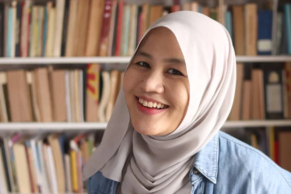 Portrait of cheerful Asian muslim female librarian wearing hijab, looking at camera and smiling, woman standing against books in library