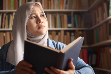 Asyalı Müslüman kadın kitap okurken düşünüyor, eğitim konsepti, kütüphanede öğreniyor, mutlu bir gülümseme ifadesi var. 