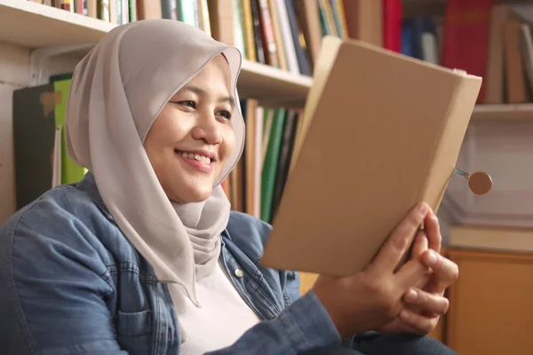 Asian muslim woman wearing hijab reading book in library, educational concept. Happy smiling expression when doing leisure activity