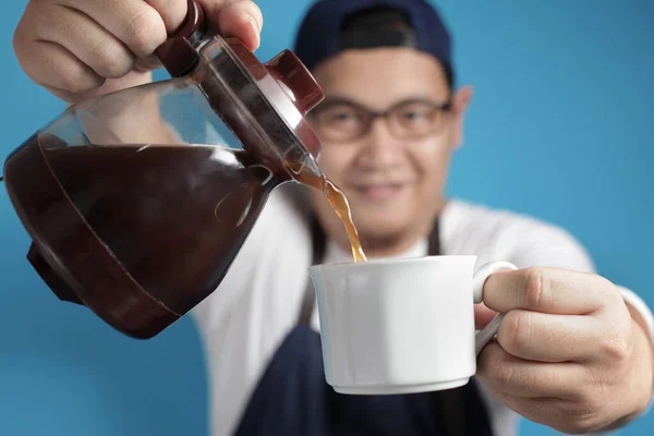 Portrait of Asian male chef or waiter smiling while pouring coffee to a cup, offering coffee concept, against blue background