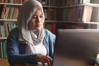 Asyalı Müslüman kadın kütüphanede okuyor, sınav hazırlığı konsepti. İnternette araştırma yapan ve laptop kullanan bir üniversite öğrencisi.