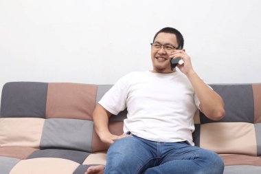 Genç gülümseyen yakışıklı Asyalı adam koltukta oturmuş telefonla konuşuyor, mobil iletişim konsepti.