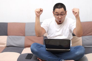 Asyalı adam zaferini kutluyor. Dizüstü bilgisayara bakıp yumruklarını sallıyor ve neşeli bir şekilde bağırıyor. Serbest çalışan rahat koltukta oturuyor.