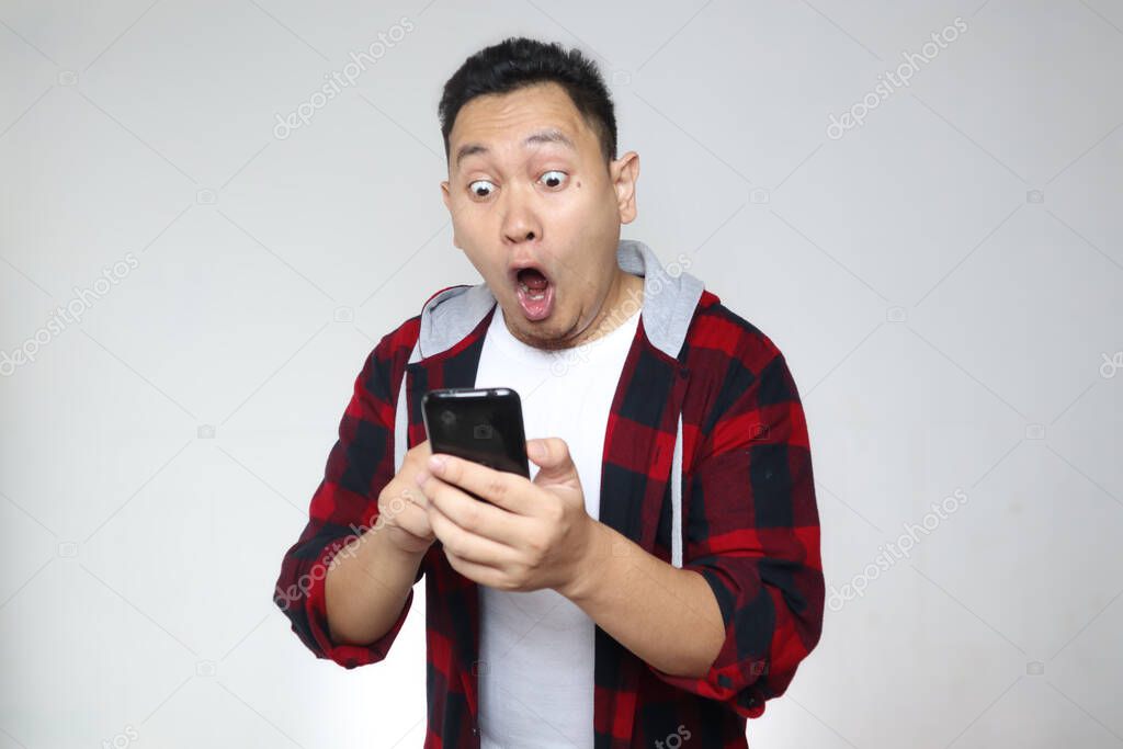 Attractive young Asian man reading texting chatting  on his phone, shocked surprised expression