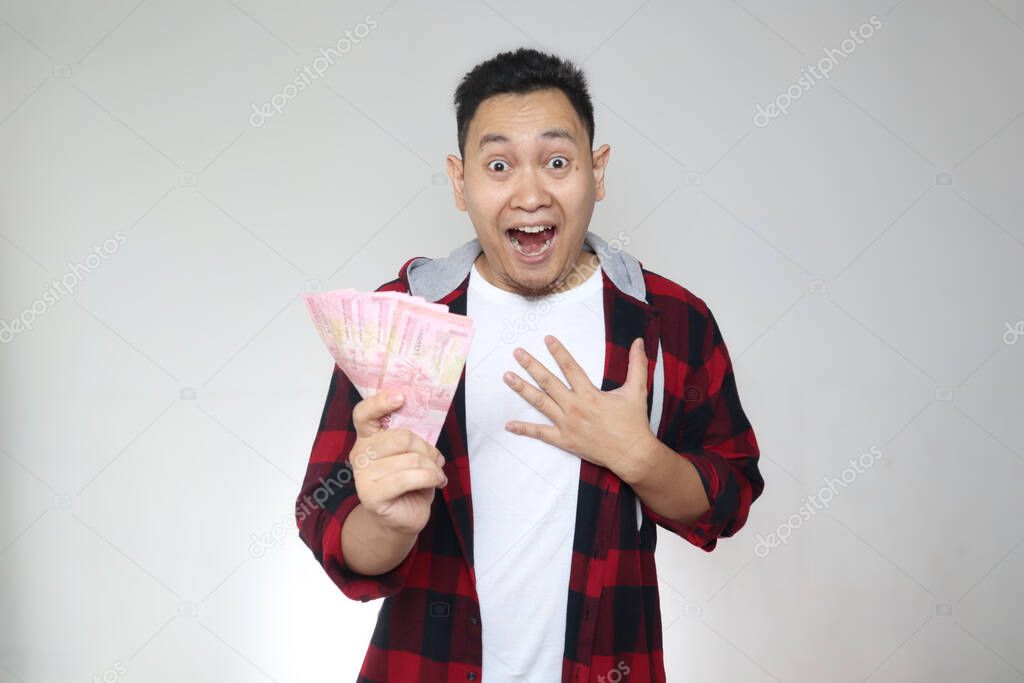 Portrait of happy young Indonesian man holding rupiah money, smiling laughing winning gesture, over white background
