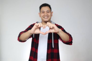 Beyaz arka planda el hareketleriyle aşk işareti gösterirken mutlu bir şekilde gülümseyen Asyalı genç adamın portresi.