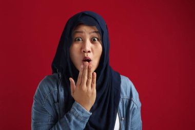 Başörtüsü takan Asyalı Müslüman kadın şaşırmış ya da şok olmuş bir ifade sergiliyor. Ağzı açık, yüzü kırmızı arkaplan üzerine kapatılmış.