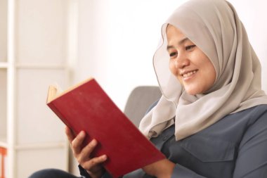 Güzel Asyalı Müslüman kadın kanepede otururken kitap okuyor, kız boş zamanlarında eğleniyor, neşeli bir ifade takınıyor.