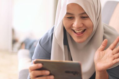 Güzel Asyalı Müslüman kadın telefonda selfie portresi yapıyor ya da kanepede otururken video çekiyor. Mutlu bir gülümseme, el sallama hareketi.