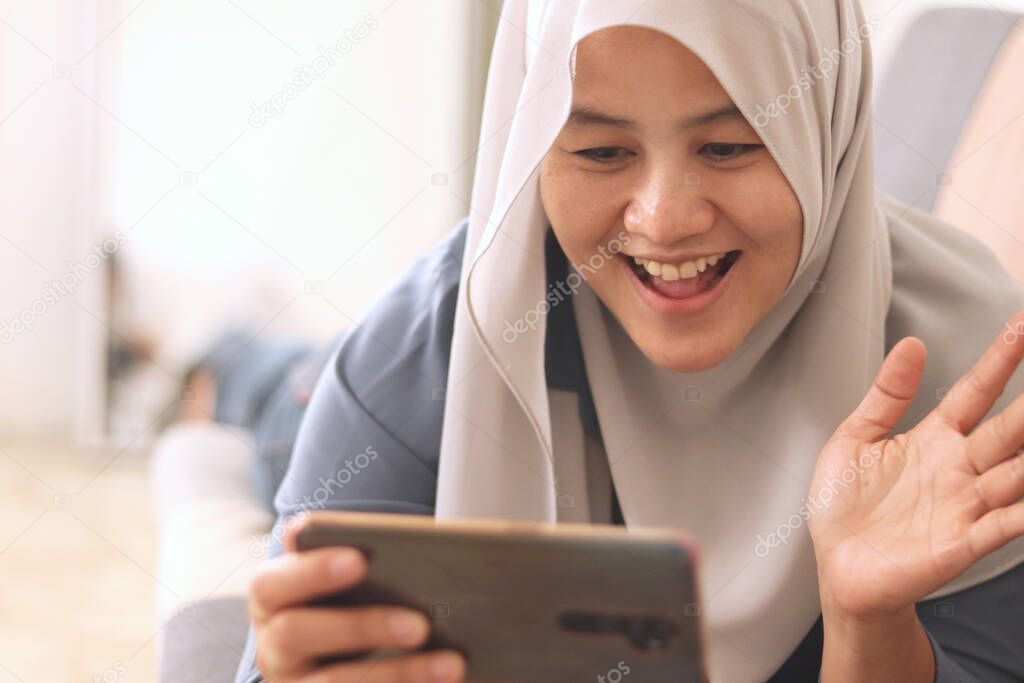 Beautiful Asian muslim lady doing selfie portrait on phone or doing video call while sitting on sofa, happy smiling cheerful expression, waving gesture