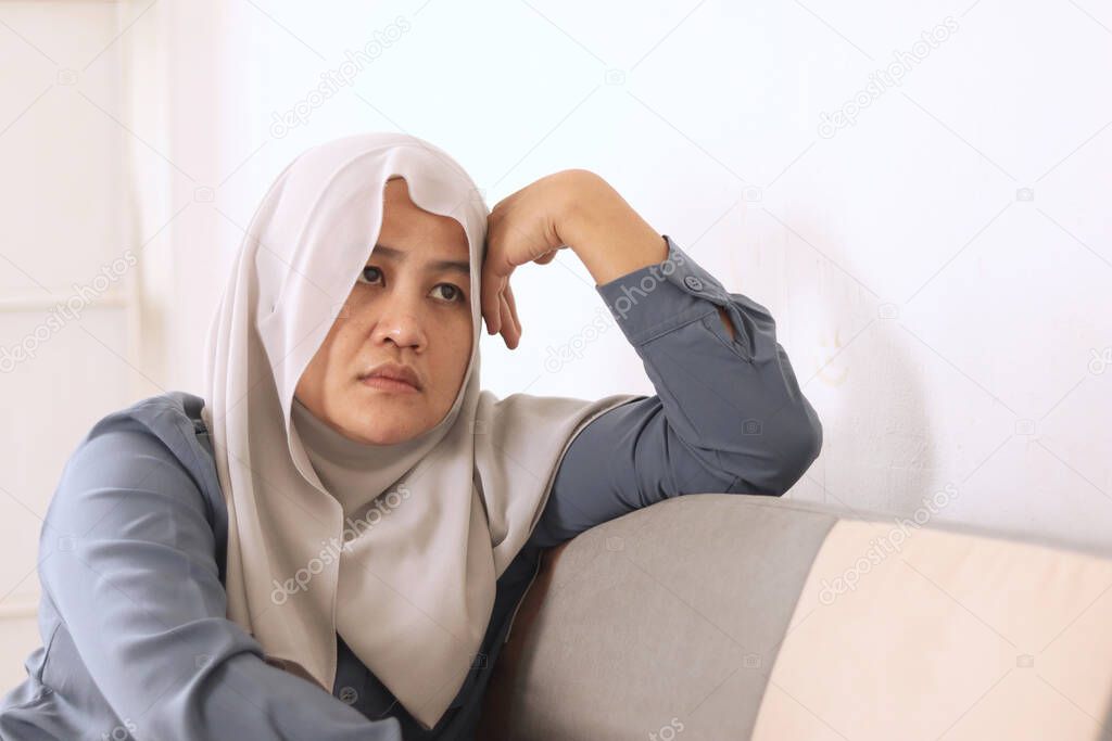 Depressed young Asian muslim woman thinking about her problems while sitting on the sofa at home
