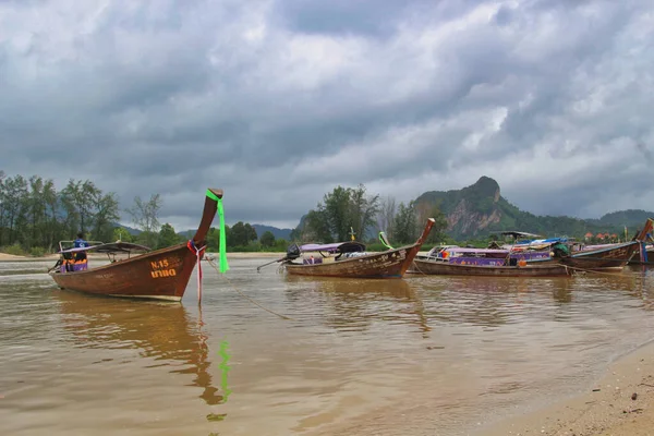 Geleneksel uzun kuyruklu tekneler Krabi 'de demirlemiş, bulutlu bir günde tropikal bir körfeze demirlemiş renkli uzun kuyruklu botlar, yemyeşil tepeler ve arka planda bir rıhtım..