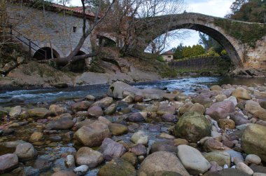 Kayalık bir nehrin üzerindeki tarihi ortaçağ köprüsü, kayalar ve kayalarla dolu sakin bir nehri kaplayan büyük bir kemerli tarihi bir taş köprünün alçak açılı görüntüsü..