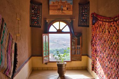 Manzaralı Fas Odası - Geleneksel bir Fas odası içinde bitki ve halılar, büyük bir pencereden tarihi bir köy manzarası sunuyor..