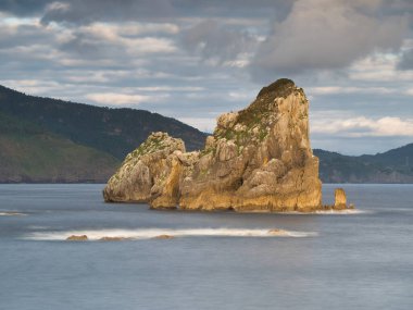 Sakin bir Bask sahil şeridinde kayalık bir deniz yığını. Denizin ortasında pürüzsüz su ve arka planda yuvarlanan dağlarla büyük bir kaya oluşumunun uzun bir görüntüsü..