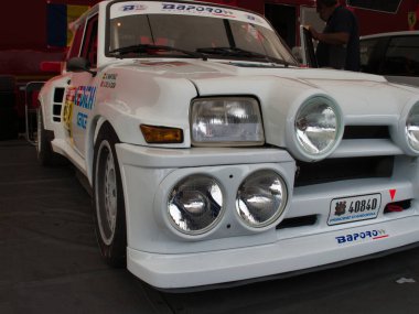 Vintage Alpine Ralli Arabası 'nın önünde beyaz bir Renault 5 Turbo yarış arabasının yakın açılı bir görüntüsü ön ızgarasını, farlarını ve çok sayıda yardımcı rali ışığını vurguluyor..