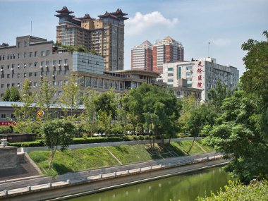 Modern gökdelenler arasındaki eski tarz binalar - yeşil ağaçlar ve çimlerle kaplı bir kanal manzarası, geleneksel Çin tarzı binalar ve arka planda modern yüksek binaların karışımı, kentsel bir ortamda eski ve yenilerin karışımını gösteriyor.
