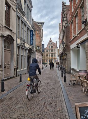 Cobblestone Ghent Caddesi 'ndeki bisikletçi - Ghent' te dar, kaldırımlı bir caddede bisiklet sürüyor, bulutlu bir gökyüzünün altında geleneksel Flaman binalarıyla dolu.