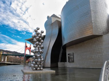 Guggenheim Müzesi Bilbao ve modern sanat. Guggenheim Müzesi ve Bilbao 'daki Nervin Nehri' nin yanında La Salve Köprüsü 'nün arka planında bulutlu mavi gökyüzünün altında yer alan modern bir heykel..