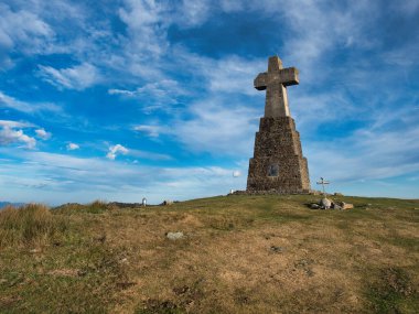 Trail Signs on a Grassy Hill - A wooden trail signpost stands on a grassy, sunlit hill with a rocky cross at the summit, guiding hikers under a wide, blue sky with wispy clouds.