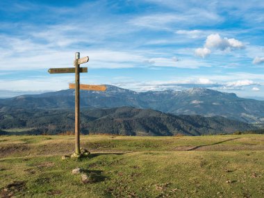 Trail Signs on a Grassy Hill - A wooden trail signpost stands on a grassy, sunlit hill with a rocky cross at the summit, guiding hikers under a wide, blue sky with wispy clouds.