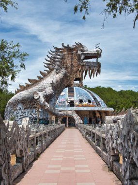 Terk edilmiş bir su parkında yıkılmış ejderha kafası, yıpranmış beton bir ejderha heykelinin yakın çekimi, açık bir ağızla bulutlu parlak mavi bir gökyüzüne karşı..