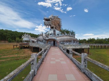 Terk edilmiş bir su parkında yıkılmış ejderha kafası, yıpranmış beton bir ejderha heykelinin yakın çekimi, açık bir ağızla bulutlu parlak mavi bir gökyüzüne karşı..