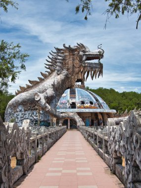 Terk edilmiş bir su parkında yıkılmış ejderha kafası, yıpranmış beton bir ejderha heykelinin yakın çekimi, açık bir ağızla bulutlu parlak mavi bir gökyüzüne karşı..