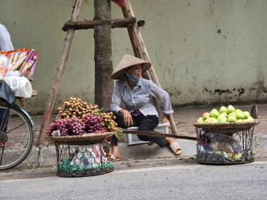 Sokak satıcısı sepetlerden taze meyve satıyor. Konik şapka giyen bir kadın Vietnam 'da kaldırımda duran tahta bir direkte dengelenmiş iki büyük sepetten taze meyve satıyor..