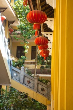 Avluda asılı parlak kırmızı fenerlerden oluşan bir sıra parlak sarı bir binanın dikey görüntüsü süslü bir merdiven ve tavandan sarkan bir dizi parlak kırmızı fener..