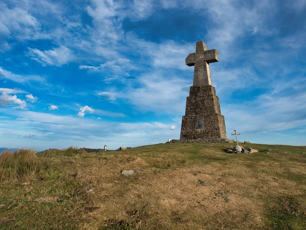 Trail Signs on a Grassy Hill - A wooden trail signpost stands on a grassy, sunlit hill with a rocky cross at the summit, guiding hikers under a wide, blue sky with wispy clouds.