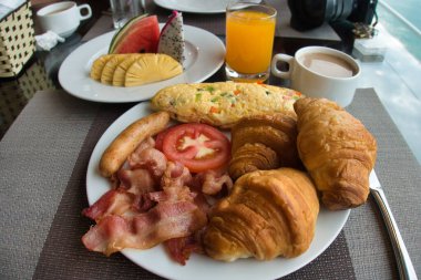 Abundant buffet breakfast with bacon and eggs - A large plate filled with bacon, sausages, eggs, and croissants, with a side of fresh fruit and a glass of juice.