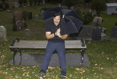 Latin bey bir mezarlığında bir bankta otururken bir şemsiye tutan. 