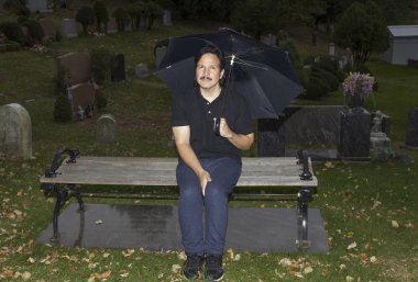 Bankta oturan ve mezarlıkta şemsiye tutan bir İspanyol adam.