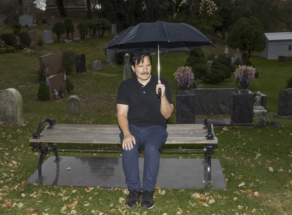 Bankta oturan ve mezarlıkta şemsiye tutan bir İspanyol adam.