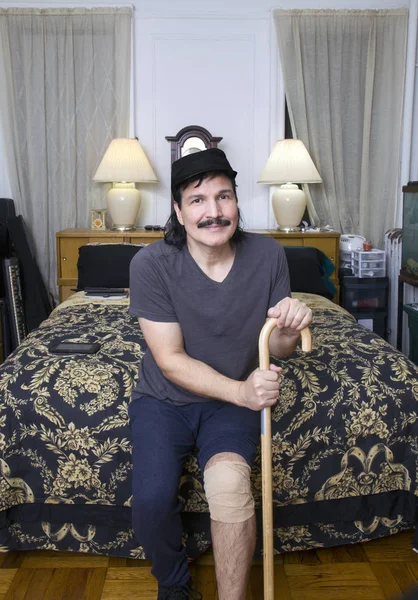 Uomo ispanico con benda al ginocchio e bastone sul letto con sorriso — Foto Stock
