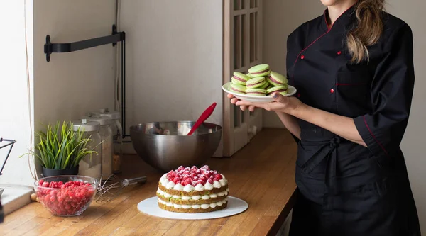 Confectioner in  Black  Work Uniform Holding Macaroons, Cake with Berries  on the Table. Kitchen Background. Confectioner, Cake, Cooking.