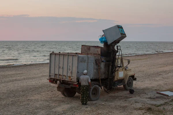 Worker of municipal recycling garbage collector truck loading waste and trash bin at Sunset. Sea on a background